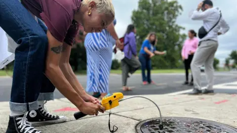 Helen Gebregiorgis A woman holding a yellow testing device measures gases emitted from street drain